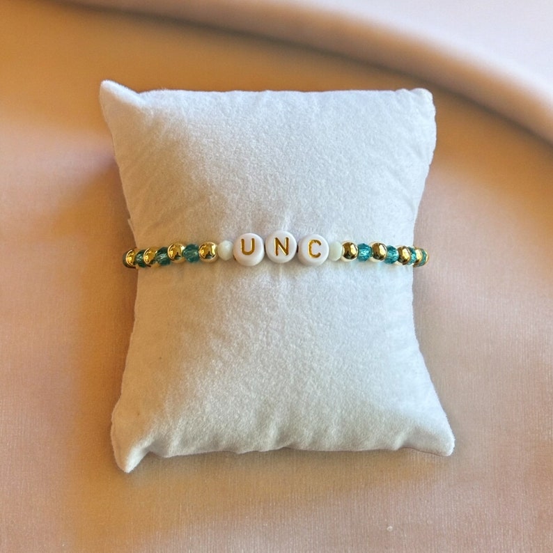 May include: A white beaded bracelet with gold accents and the letters "UNC" spelled out in gold lettering.