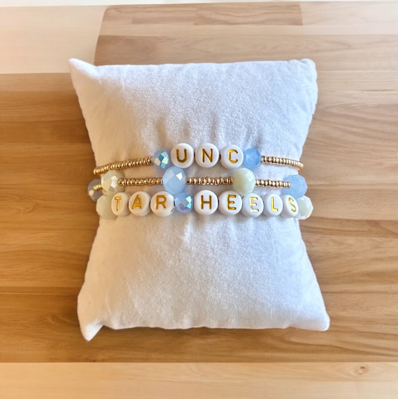 May include: Three beaded bracelets with gold accents spelling out "UNC" and "TAR HEELS" in white letters. The bracelets are on a white pillow.