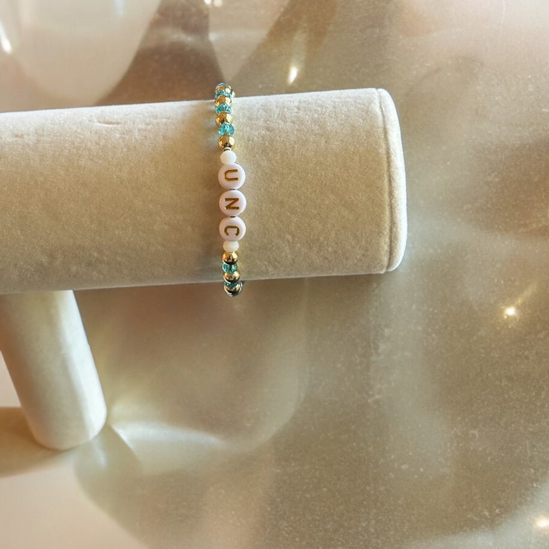 May include: A gold and turquoise beaded bracelet with white letter beads spelling out "UNC".