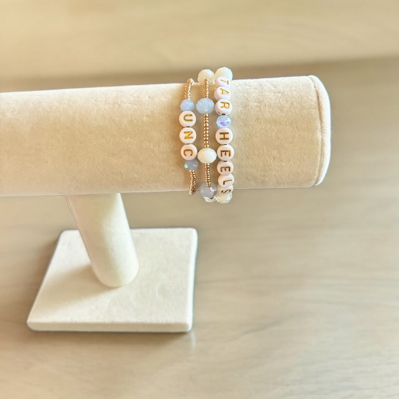 May include: Three beaded bracelets with gold lettering. The bracelets spell out "UNC", "TAR", and "HEELS".