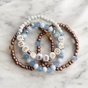 May include: Four beaded bracelets stacked together. The bracelets feature white, light blue, and bronze-colored beads. One bracelet spells out "THE CITADEL" with gold-colored letter beads. The bracelets are arranged on a white and gray marble surface.