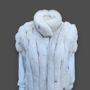 Puede incluir: Un chaleco de piel blanco con cuello alto. El chaleco está hecho de piel suave y esponjosa y tiene una cremallera.
