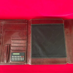Puede incluir: Una carpeta de cuero marrón con una calculadora incorporada. La carpeta tiene varios bolsillos y un cierre de cremallera.