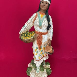 May include: A ceramic figurine of a Native American woman wearing a white dress with orange and blue accents. She is holding a basket of fruit and a pitcher. The figurine is standing on a base with green grass.