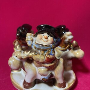 May include: A ceramic figurine featuring a snowman riding a reindeer, with three other snowmen on reindeer. The snowmen wear black hats and blue and brown outfits. The base is a cream color with gold trim.