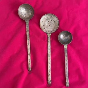 May include: Three antique metal kitchen utensils: a ladle, a slotted spoon, and a smaller ladle. The utensils have long handles with decorative accents and a hammered texture on the bowls. The background is a solid red color.