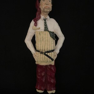 May include: A ceramic figurine of a person in a white shirt, green and white striped apron, and burgundy pants. The figure has a black belt, a green polka-dot tie, and a red head covering. The figurine is standing with hands on hips.