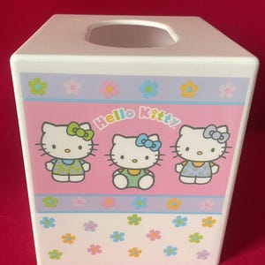 May include: White plastic tissue box cover with a pink background and three Hello Kitty characters. The characters are wearing different colored outfits and have bows in their hair. The background has a purple and blue border with a row of colorful flowers. The bottom of the box has a white background with a scattered pattern of colorful flowers.