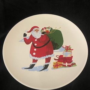 May include: A round, off-white plate featuring a festive Santa Claus illustration. Santa is depicted in a red suit with a white beard, carrying a green sack of gifts. Several wrapped presents are stacked nearby, creating a holiday theme.