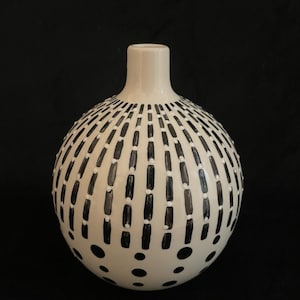 May include: A cream-colored, spherical vase with a narrow neck. The vase is decorated with a pattern of black, vertical dashes and scattered black dots. The design covers the majority of the vase's surface, creating a striking contrast against the cream background.