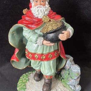 May include: A figurine of Santa Claus dressed in green and red attire, holding a black pot overflowing with gold coins. The figure stands on a base with a stone and grassy area. The Santa figure has a white beard and a red hat.