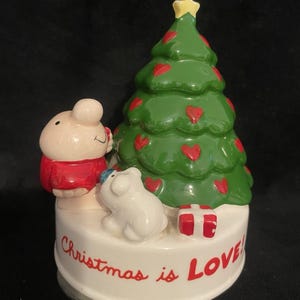 May include: A ceramic Christmas decoration featuring a green Christmas tree with red heart ornaments and a yellow star. Two small animal figures and a gift box are on a white base with the words "Christmas is Love!" in red.