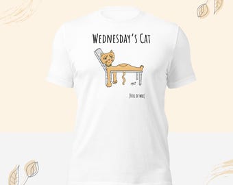 Könnte beinhalten: Ein weißes T-Shirt mit kurzen Ärmeln und einer Grafik einer orangefarbenen Katze, die auf einem Stuhl liegt. Der Text über der Katze lautet "WEDNESDAY'S CAT" und darunter steht "(FULL OF WOE)". Die Katze schaut traurig und trägt ein grünes Halsband.