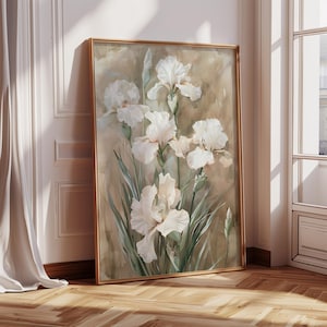 May include: A framed art print featuring a watercolor painting of white iris flowers with green leaves against a muted beige background. The artwork is displayed in a gold-colored frame, leaning against a wall with a wooden floor.