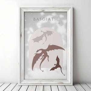 May include: A white framed print with the word "BASGIATH" in pink lettering. The print features three silhouettes of dragons in shades of pink and brown against a white background.