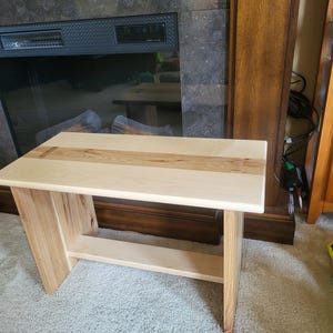 Peut inclure: Un banc en bois clair avec un dessus rectangulaire et une étagère inférieure. Le banc a une finition en grain de bois naturel avec une bande de bois plus claire sur le dessus. Les pieds sont inclinés, offrant une stabilité.