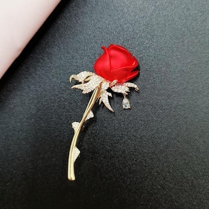 May include: A red rose brooch with a gold stem and leaves, embellished with sparkling crystals. The brooch has a teardrop-shaped crystal pendant. The rose is set against a dark background.