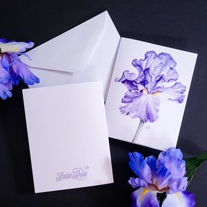 May include: A set of blank greeting cards and envelopes featuring a watercolor painting of a purple iris flower. The cards are white, with the artist's signature in the lower right corner. The back of one card has the artist's logo.