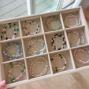 Può includere: Un vassoio portagioie beige con dodici scomparti, ognuno contenente un braccialetto di perline unico. I braccialetti presentano vari design, tra cui piccole perline bianche, verdi, blu, rosa e nere, alcune con accenti di perle e motivi floreali.