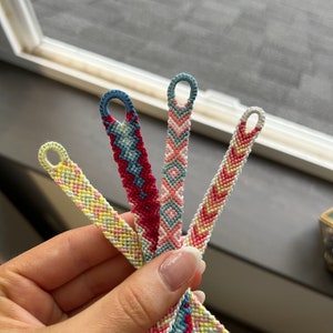 Puede incluir: Cuatro pulseras de la amistad hechas a mano en varios colores, incluyendo amarillo, rosa, rojo y azul. Cada pulsera presenta un patrón tejido y un lazo en el extremo. Las pulseras se sostienen en una mano, mostrando sus intrincados diseños.