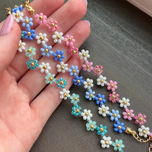 May include: Several beaded daisy chain bracelets in shades of blue, pink, white, and teal. Each bracelet features small, colorful flower beads with gold-colored centers and chain clasps. The bracelets are displayed on a neutral surface.
