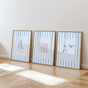 May include: Three framed watercolor prints with a beach theme. The prints feature a sailboat, surfboards, and a bikini hanging on a clothesline. The frames are a light brown color, and the background of the prints has blue and white stripes.