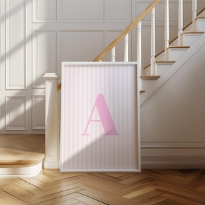 May include: A framed print featuring a large pink letter "A" against a background of vertical pink and white stripes. The print is displayed in a white frame and leans against a wall near a staircase with wooden steps and a white banister.