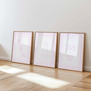 May include: Three framed art prints with a beach theme. Each print has a white background with a pink border and a pink illustration. The illustrations include seashells, a sailboat, and a bikini.