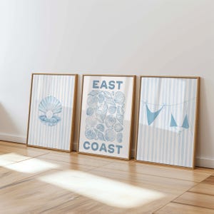 May include: Three framed art prints with a coastal theme. The prints feature a seashell, a collection of seashells with the text "EAST COAST", and a blue bikini hanging on a clothesline. Each print has a light blue and white striped background.