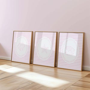 Puede incluir: Tres impresiones artísticas enmarcadas con diseños marinos. Cada impresión presenta una concha, una estrella de mar o una concha de vieira sobre un fondo a rayas rosas y blancas, dentro de un marco ovalado blanco. Los marcos son de color marrón claro.
