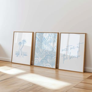 May include: Three framed art prints with beach-themed designs. One features a palm tree and surfboard, another a blue seashell pattern, and the third reads "meet me at the beach" in script. The frames are a light brown color.