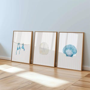 May include: Three framed art prints with a beach theme. The prints feature watercolor illustrations of a blue bikini hanging on a clothesline, a sand dollar, and a blue seashell. The frames are a light brown color, and the background is a striped pattern.