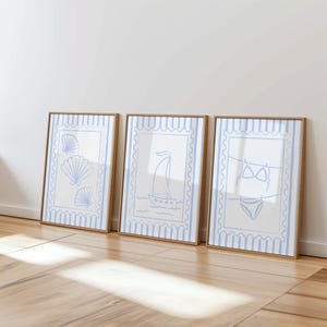 May include: Three framed art prints with a coastal theme. Each print features a white background with a light blue border and design. The designs include seashells, a sailboat, and a bikini. The frames are a light brown color.