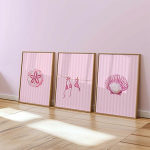 May include: Three framed art prints with a beach theme. The prints feature a pink sand dollar, a pink bikini hanging on a clothesline, and a pink seashell. Each print has a pink and white striped background and a wooden frame.