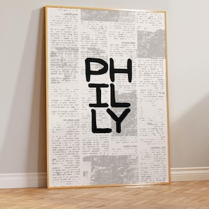 May include: A framed art print featuring the word "PHILY" in bold black letters. The background is a collage of newspaper print in shades of gray and white, with a light wood frame.