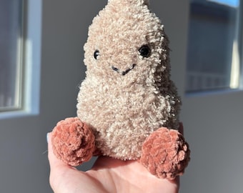 Puede incluir: Un peluche beige con una cara sonriente y dos pies redondos de color coral. El peluche tiene un aspecto texturizado y difuso. El juguete se sostiene en una mano.