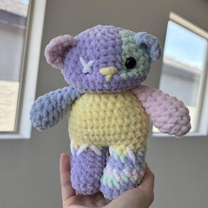 May include: A plush crochet bear toy featuring a multi-colored design with a purple and mint green head, a yellow torso, and mismatched limbs in blue, pink, and a variegated pastel yarn. The bear has a white stitched X for one eye and a black bead eye.
