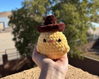 May include: A yellow crocheted chick wearing a brown sombrero. The chick has black eyes and a small orange beak.