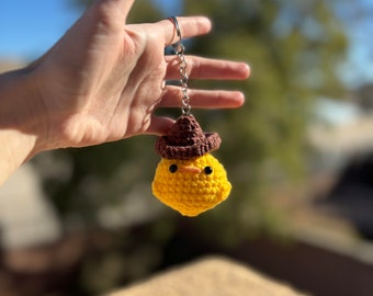 May include: A yellow crocheted chick wearing a brown crocheted hat, attached to a silver keychain.