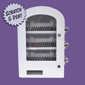 May include: White vending machine with a clear front, three shelves, and a coin slot. The machine has a silver handle and the text "SCRATCH & DENT" in a starburst graphic.