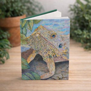 May include: A hardcover journal featuring a detailed illustration of a bearded dragon. The artwork showcases the reptile's textured skin, blue eye, and the surrounding foliage. The journal has a green spine and blank pages.