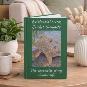 May include: A hardcover book with a green cover, titled "Existential crisis, Cricket thoughts." The cover features an illustration of a lizard. The book is on a wooden table with plants and a vase in the background.