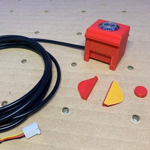 May include: A red 3D printed box with a blue lens on top, connected to a black wire and a white connector. The box is accompanied by two red and yellow pieces and a red button.