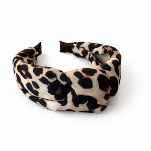 Könnte beinhalten: Ein geknotetes Stirnband mit Leopardenmuster. Das Stirnband hat einen beigefarbenen Hintergrund mit schwarzen und braunen Flecken. Die Enden des Stirnbandes sind einfarbig braun. Ein Haaraccessoire.