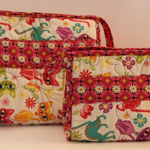 May include: Two quilted zippered pouches with a white background and a pink, black, and white floral pattern. The pouches feature a whimsical design of cats with colorful accents.
