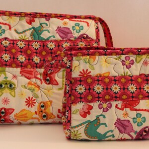 May include: Two rectangular quilted pouches of different sizes featuring a white background with a colorful pattern of stylized cats, flowers, and abstract shapes. Each pouch has a horizontal band with a pink and orange floral print.