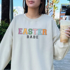 Może przedstawiać: Kremowa bluza z napisem "EASTER BABE" w kolorach tęczy.