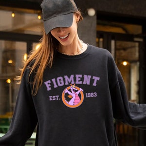 Puede incluir: Una sudadera negra con un gráfico morado y rosa de un personaje de dibujos animados. La sudadera tiene el texto "FIGMENT EST. 1983" impreso en la parte delantera.