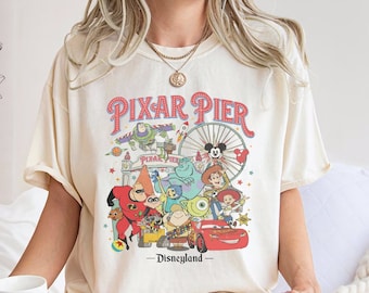Może przedstawiać: Biała koszulka z grafiką w stylu vintage przedstawiającą Pixar Pier w Disneylandzie. Grafika przedstawia postacie z popularnych filmów Pixara, w tym Buzz Astral, Woody, Mike Wazowski, Sulley i Pan Niesamowity. Tekst "Pixar Pier - Disneyland" jest nadrukowany pod grafiką.
