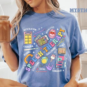 May include: A light blue t-shirt with a colorful graphic design that says "Speech Therapy" and includes illustrations of speech therapy tools and concepts. The design includes words like "Articulation", "Cognition", "Hearing", "Modalities", "Swallowing", "Voice", "Stuttering", "Social", and "Language".